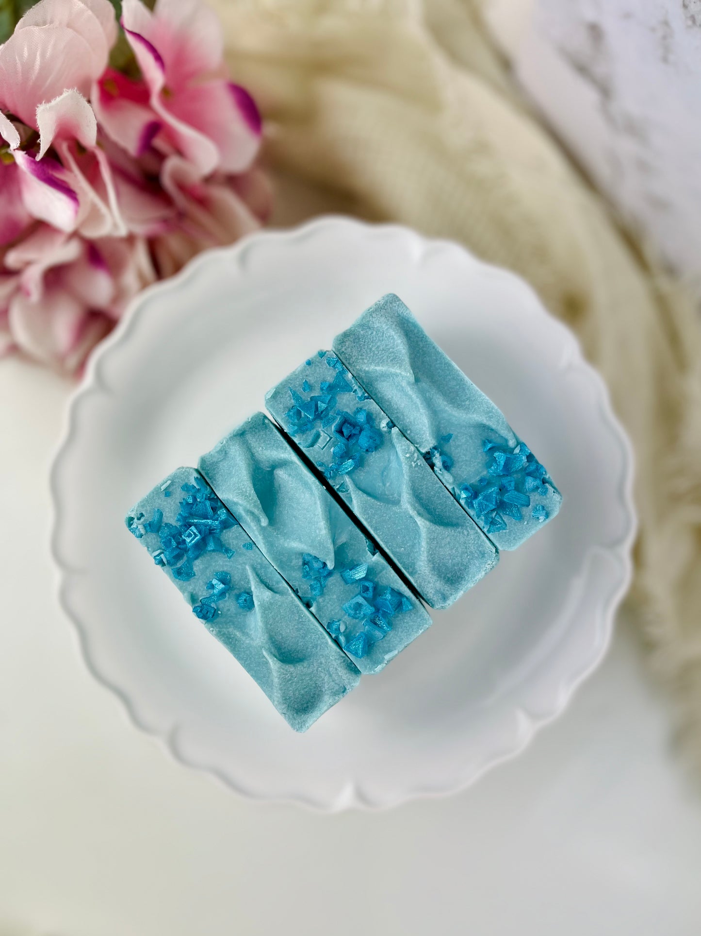 Blue soap bars with a textured surface on a white plate, with pink flowers in the background.
