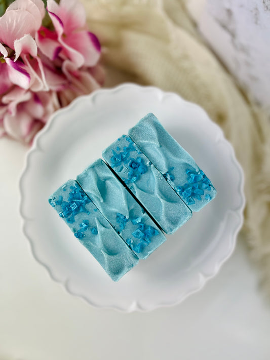 Blue soap bars with a textured surface on a white plate, with pink flowers in the background.